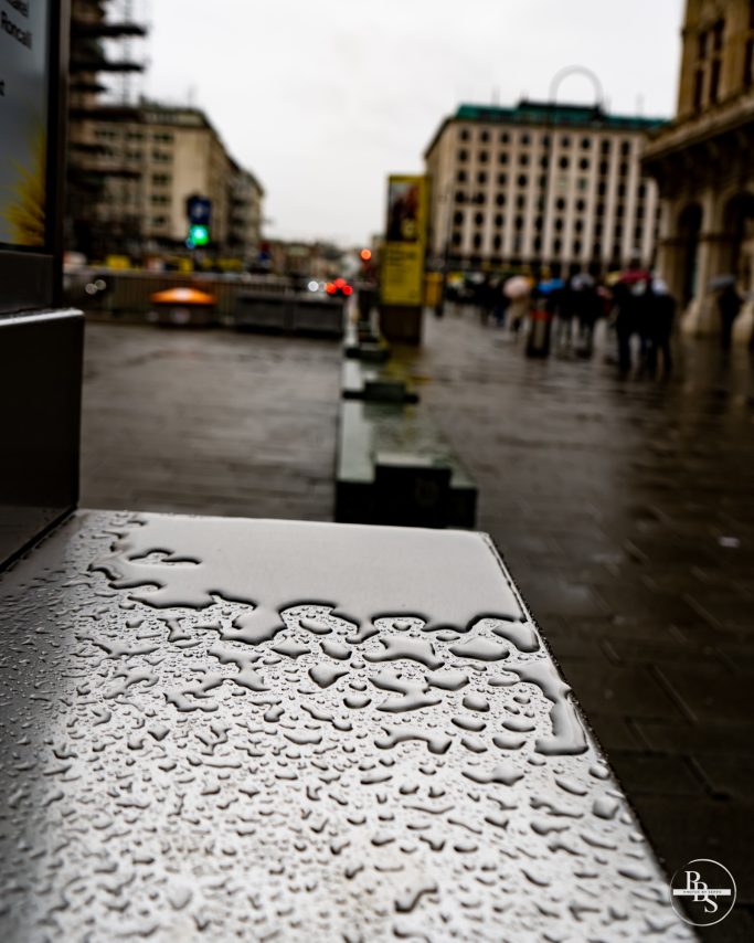 Nasse Stadtstraße mit Wasserpfützen und Menschen im Hintergrund.