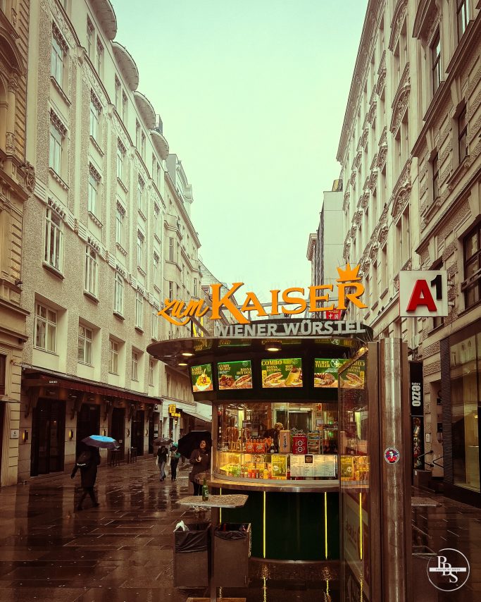 Kiosk mit orangefarbenem "Kaiser"-Schild in einer regnerischen Stadtstraße.