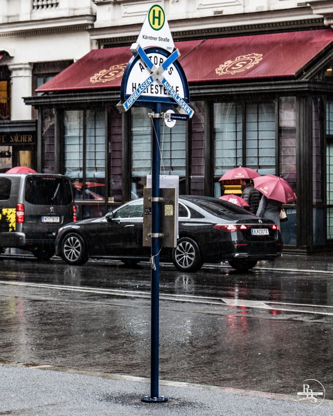 Haltestelle mit Regenschirm-bedeckten Passagieren und Regen auf der Straße.