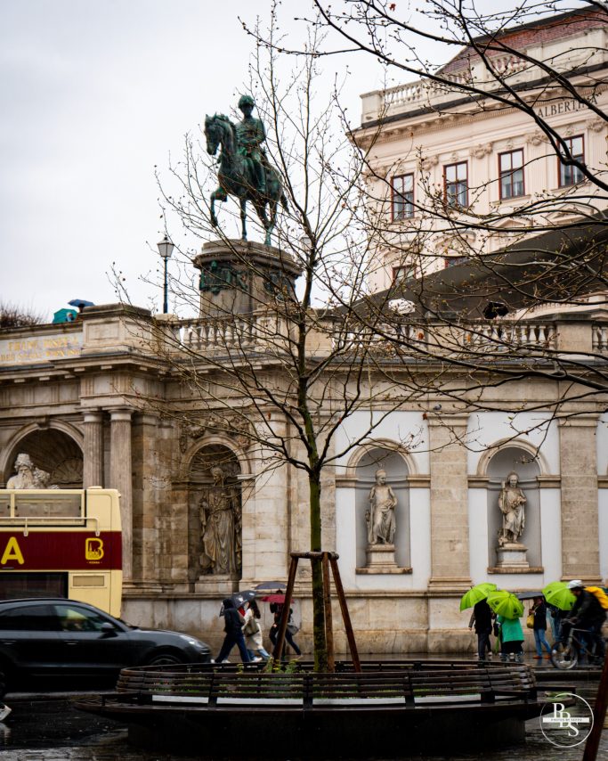 Reiterstatue vor historischer Gebäudefassade, umgeben von Bäumen und Passanten.