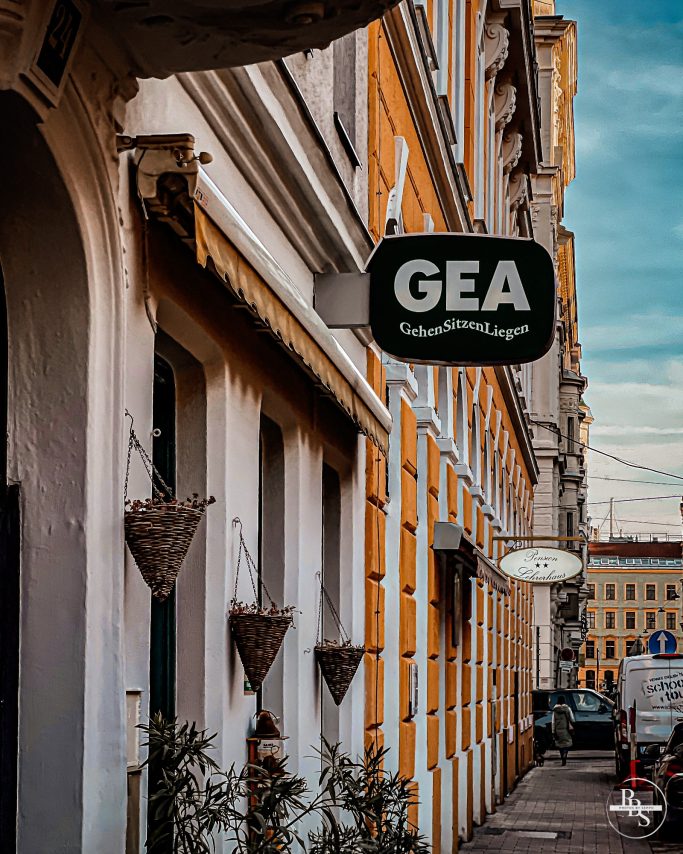 Gelbe Fassade eines Gebäudes mit einem schwarzen Schild "GEA" und hängenden Pflanzen.