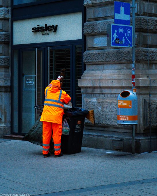 Mitarbeiter in orangefarbener Kleidung leert einen Abfallbehälter auf der Straße.